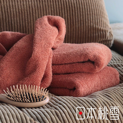 一堆粉紅色毛巾堆疊在燈芯絨質感的沙發上，一旁擺放著木柄梳子，右下角有「日本桃雪」的文字標記。