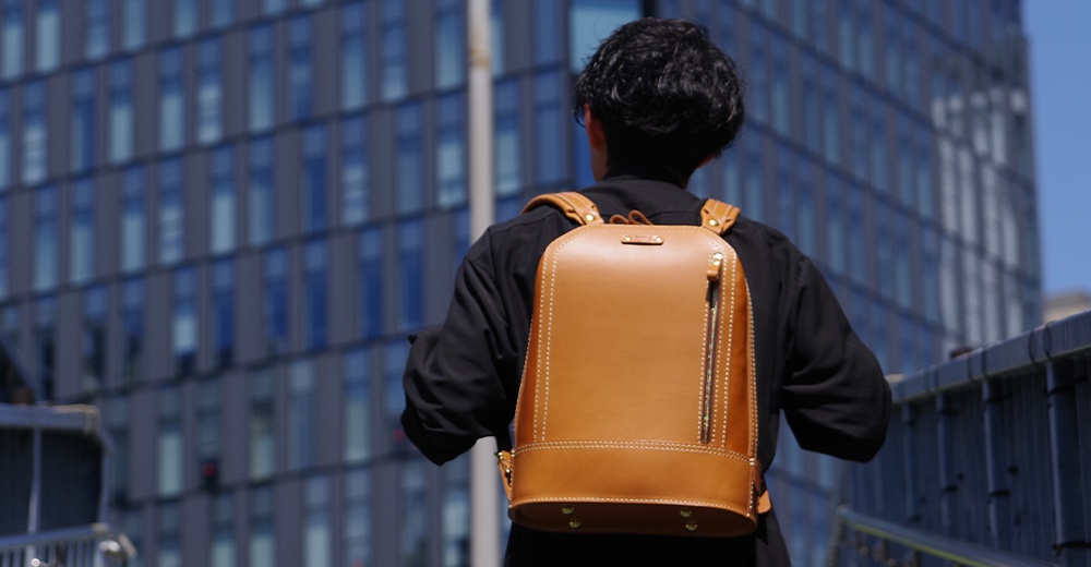 皮革立體商務後背包，採用棕褐色皮革製成，設有拉鍊主隔層和縫線細節，搭配同色系皮革肩帶。