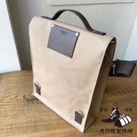 米色純棉帆布製成，帶有皮革提把及側肩背帶，正面有深棕色皮革方形裝飾片，並印有品牌標誌。