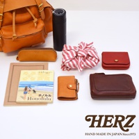 職人手作皮革復古風旅行後背包 R-65，包含一個橘棕色皮革後背包、一個橘色筆袋、一個黑色保溫瓶、一條紅白色條紋布包、一個紅色皮夾、一本 Honolulu 圖書、一個橘色皮夾和一個深棕色皮夾。