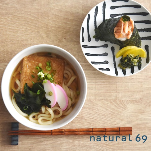 白瓷丼碗盛著日式烏龍麵，配料有炸豆腐、海帶、魚板，旁為裝有飯糰、鮭魚、漬物及海苔的設計風盤，木紋桌面擺放一雙竹筷。
