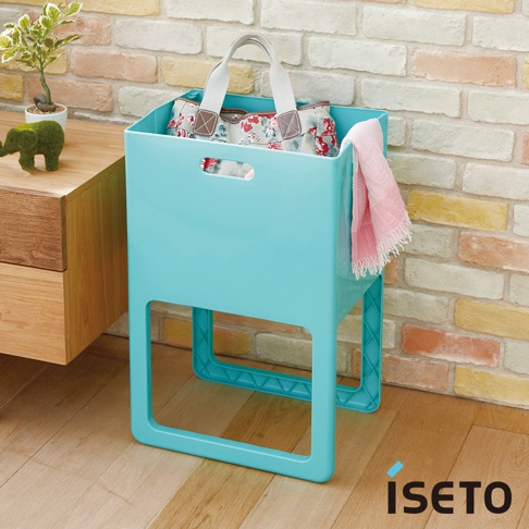 一個淺藍色的折疊高腳置物籃，籃內裝有一個印有花卉圖案的袋子，籃子側面掛著一條粉色毛巾，置物籃由塑膠製成，整體設計簡潔實用。
