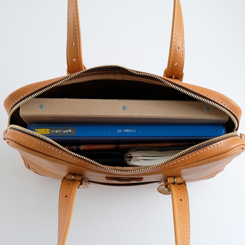 棕褐色皮革公事包，頂部拉鍊敞開，露出內部空間，可容納藍色文件夾、紙本書籍及其他物品。包包配有兩條短提把。