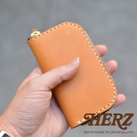 職人手作淺棕色皮革橢圓形鑰匙包，以手工縫線裝飾，下方印有HERZ品牌標誌和HAND MADE IN JAPAN Since1973字樣。