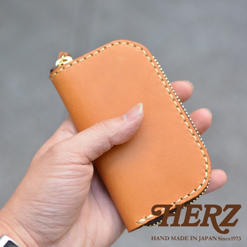 職人手作淺棕色皮革橢圓形鑰匙包，以手工縫線裝飾，下方印有HERZ品牌標誌和HAND MADE IN JAPAN Since1973字樣。