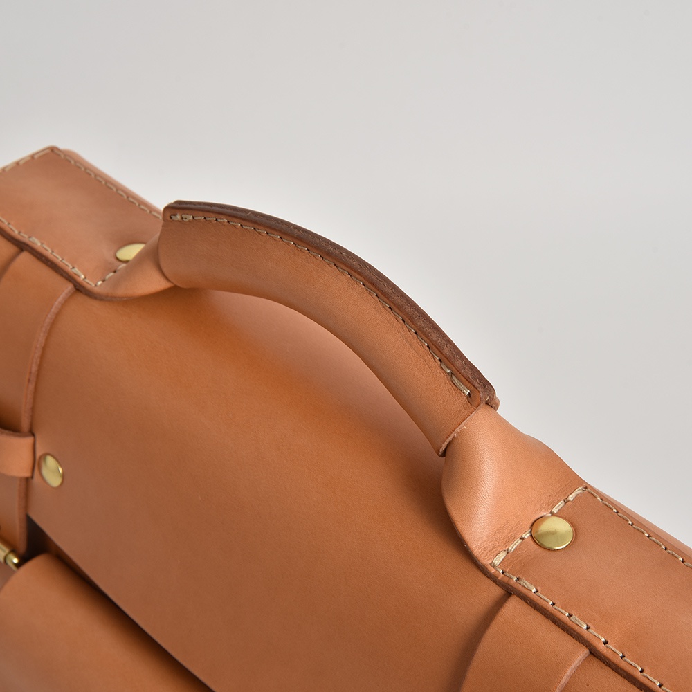 職人手作皮革學院風兩用商務公事包，近距離特寫，展示其天然皮革材質、縫線細節及金色鉚釘。提把部分為圓弧形，皮革呈現溫潤的淺棕色調。