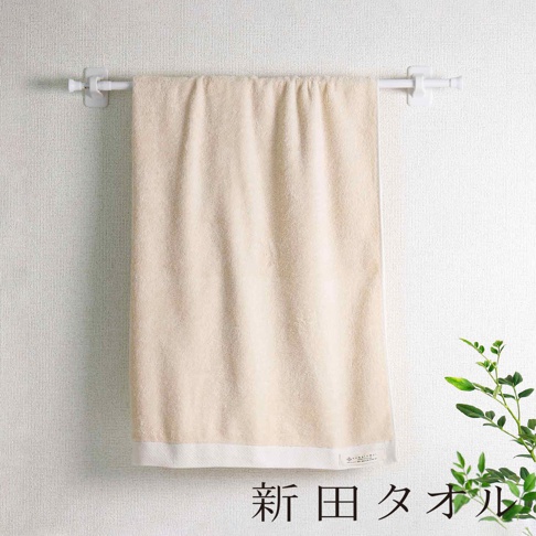 一條淺米色有機棉柔無漿生浴巾，懸掛於白色毛巾架上，浴巾紋理細膩，下緣有一條白色滾邊，背景為白色紋理牆面，右側有綠葉植物點綴。