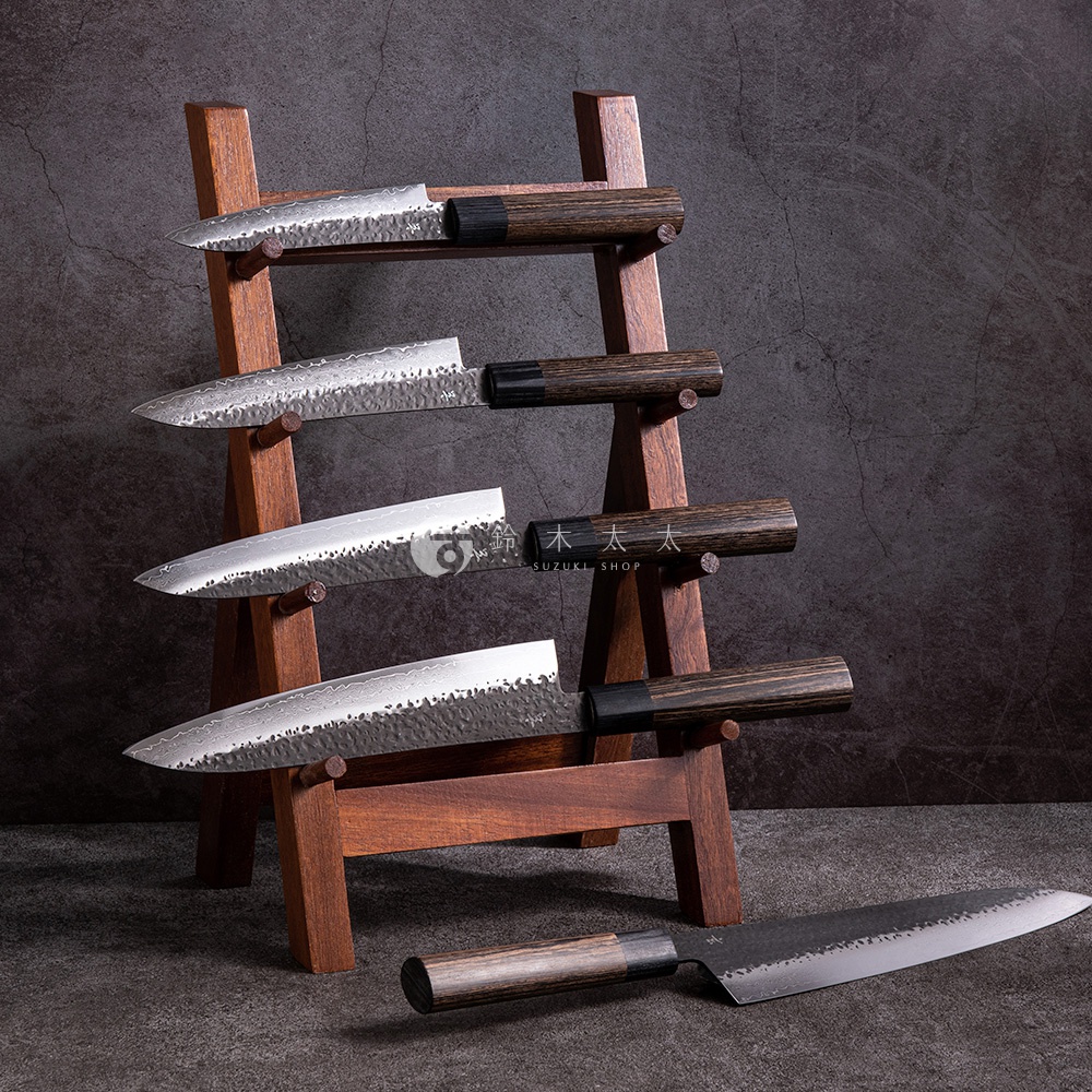 木製刀架上擺放著四把銀色金屬主廚刀，刀刃呈現錘紋和類似大馬士革鋼的紋路。刀柄為深棕色木質，搭配黑色接環。右下方另外擺放著一把相同設計的主廚刀，刀刃同樣是銀色金屬，刀柄為深棕色木質。
