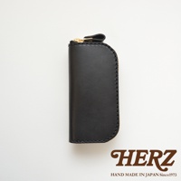 黑色皮革橢圓形鑰匙包，配有金色拉鍊和一個皮革拉環，底部印有HERZ品牌標誌。