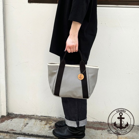 灰色帆布製手提托特包，配有黑色提把和白色內襯，包包上掛有船錨造型的木質吊飾。