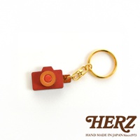 職人手作皮革相機造型鑰匙圈，紅褐色皮革機身，搭配橘色鏡頭與紅色快門按鈕，配有金色金屬掛環與鎖扣。