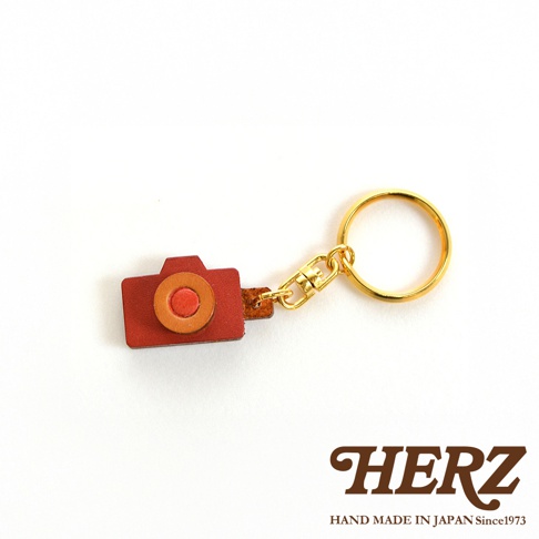 職人手作皮革相機造型鑰匙圈，紅褐色皮革機身，搭配橘色鏡頭與紅色快門按鈕，配有金色金屬掛環與鎖扣。