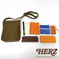 日本HERZ職人手作皮革，綠色軟皮斜背包、橘色皮夾、筆記本、藍色印花頭巾，以及各式皮革小物，搭配品牌HERZ的標誌。