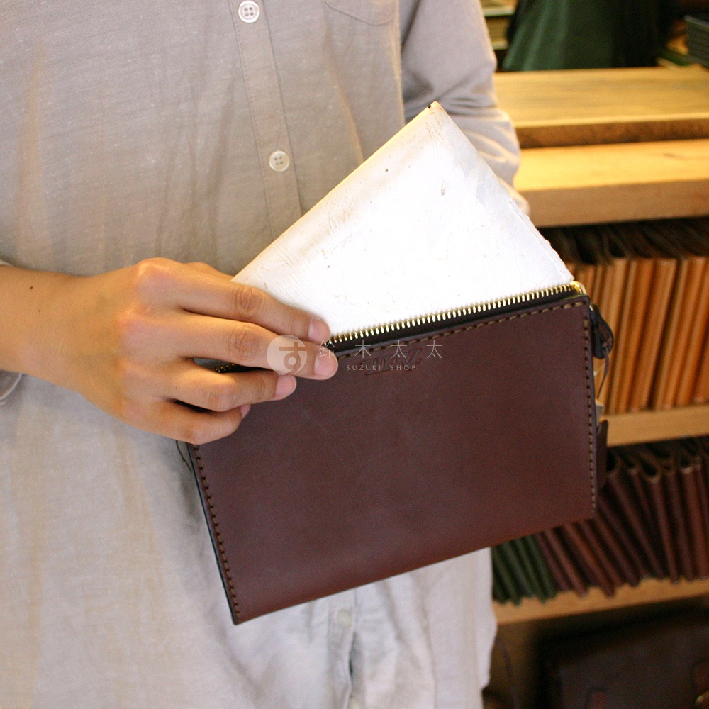 手拿包為深棕色皮革，配有金色拉鍊，側面縫線清晰可見。