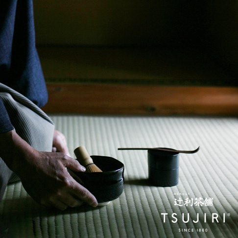 一手捧著黑色抹茶碗，碗中置有竹製抹茶茶刷，碗旁有一個黑色茶則收納盒，盒上放著一把木製茶則。