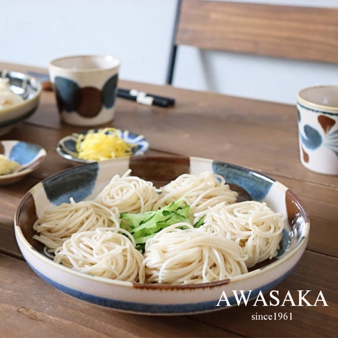 筆青釉繪料理盤盛裝著白色細麵，盤緣飾有藍色、棕色與米白色塊狀紋理。一旁還有裝有黃色絲狀配料與醬料的小碟，以及兩個帶有藍色與棕色圓點或花葉圖案的杯子。