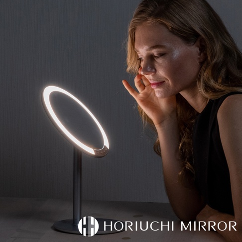 銀色金屬底座的圓形LED化妝鏡，鏡面散發著柔和的白光，一位女子正在鏡子前進行臉部保養。