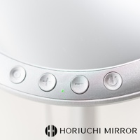 銀色桌上型化妝鏡的局部特寫，顯示鏡面邊緣的控制按鈕，包括電源、加號、減號，以及一個綠色指示燈。