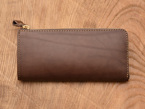 深棕色皮革L型長夾，設有雙拉鍊，搭配金屬拉鍊頭與皮革拉環，縫線清晰，材質光滑，放置於木紋背景上。