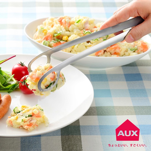 一雙銀色金屬製的leye 靈活炸物夾，夾著馬鈴薯沙拉，正要夾入白色盤子，盤上有沙拉、番茄和香腸。