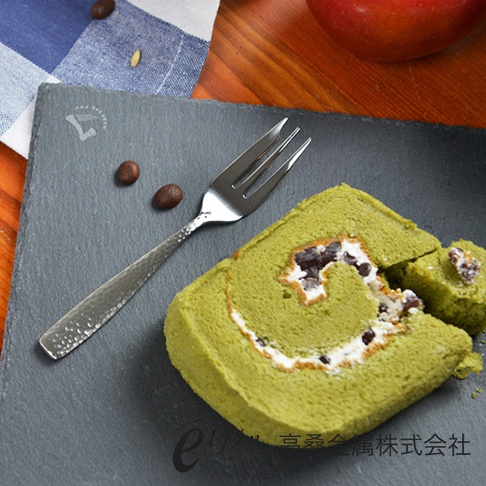 銀色不鏽鋼叉子，帶有錘目紋，置於黑色石板上，旁有一塊綠色抹茶蛋糕捲和咖啡豆。