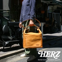 一位男士手提著一個淺棕色厚實皮革單肩波士頓包，包包上有一個方形前袋和一個小圓形品牌標誌。