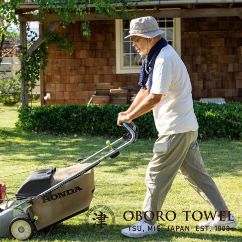 一位長者在草地上推著割草機，他身穿淺色上衣、卡其色長褲，頭戴帽子，脖子上圍著深藍色圍巾。割草機的集草袋上印有「HONDA」字樣。畫面右下角有「OBORO TOWEL」的品牌標誌。