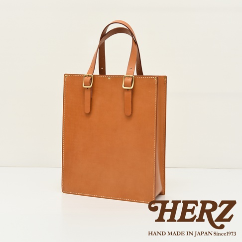 職人手作皮革商務用紙袋托特包，呈現焦糖色光滑皮革，兩側有可調節銅扣。提把設計簡約，縫線精緻，整體風格為經典都會。