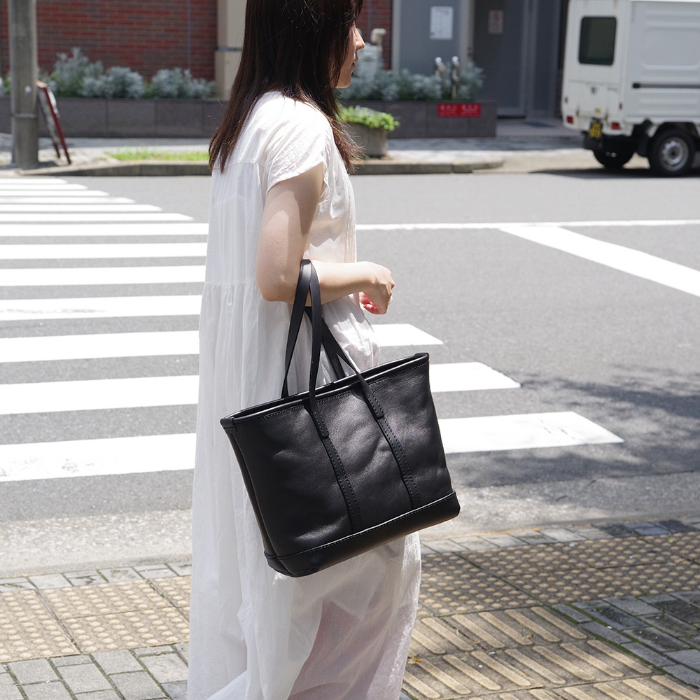 一位女性身穿白色洋裝，手提一個黑色皮革經典托特包，包包有兩個提把，皮革表面平滑，有縫線裝飾。