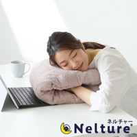 一位女子趴在淡粉色毛巾材質的甜甜圈形狀枕頭上休息，枕頭放在桌面上，旁邊有筆記型電腦和杯子。