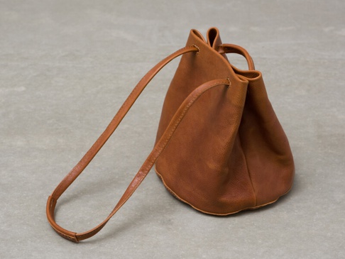 棕褐色皮革製成、抽繩開口的圓筒狀單肩包，配有可手提的短提把和一條長肩帶。