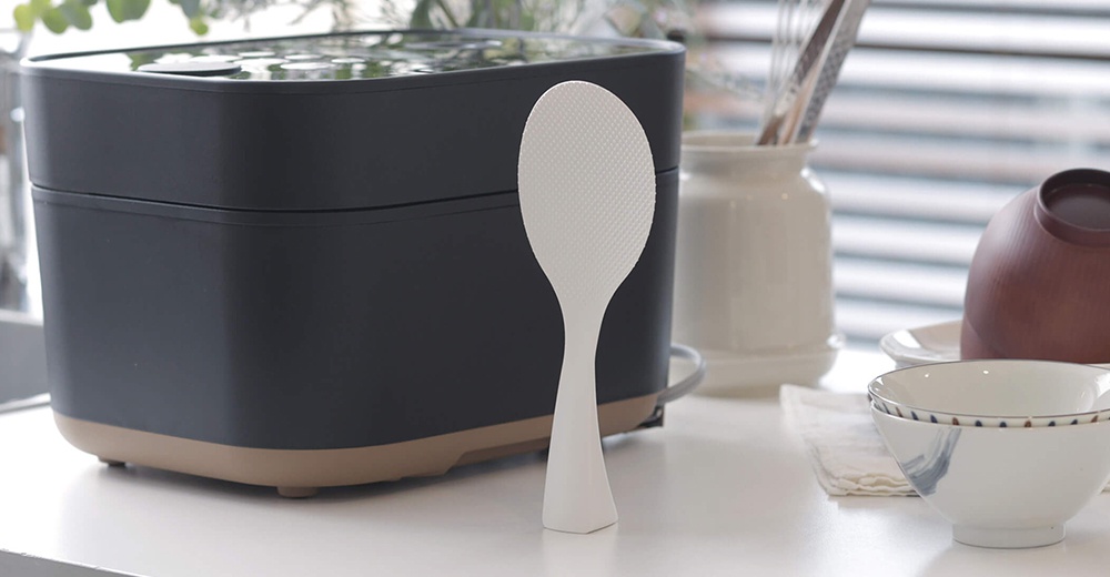 一個黑色的電鍋旁放著一個白色的站立式飯匙，後面是兩個碗和一個罐子。