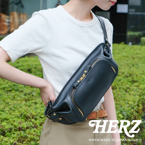 一名女子身穿白色短袖上衣，斜背一個黑色手工皮革拉鍊旅行斜背包，包包上有HERZ品牌標誌，背景是綠色植物，下方有HERZ品牌名稱及HAND MADE IN JAPAN Since1973字樣