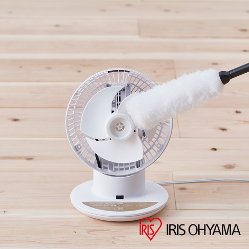 白色 PCF-SC15T 空氣循環扇，擁有簡約的圓形底座和傾斜角度可調的白色扇網，風扇中心有一個白色扇葉，一個白色毛刷正伸入扇網清潔。產品放置在淺木紋地板上，右下方有 IRIS OHYAMA 的紅色心形 Logo。