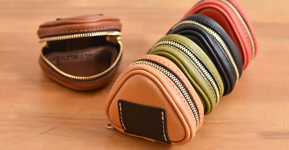 職人手作皮革小物收納包，造型為三角飯糰，共展示棕色、橘色、綠色、黑色、紅色五種顏色，皆配有金色拉鍊。