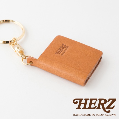 職人手作皮革書本造型鑰匙圈，淺棕色皮革，附有金色金屬鑰匙圈和扣環，皮革表面壓印有「HERZ MADE IN JAPAN」字樣。