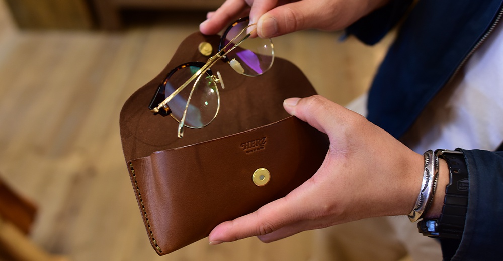 一雙圓框眼鏡正在被放入一個棕色皮革眼鏡盒中，盒子上印有HERZ的壓印字樣。