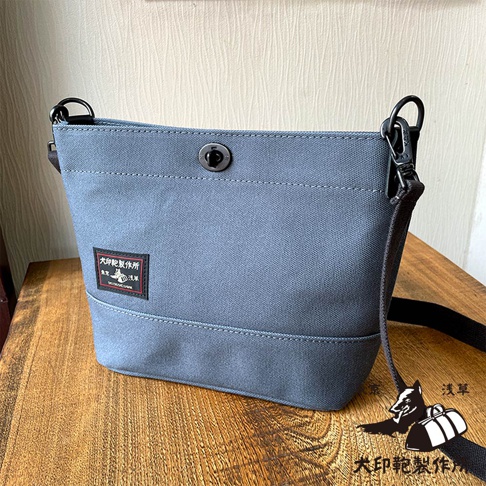 犬印鞄製作所的灰色純綿帆布迷你側背包，帶有品牌標籤和金屬鎖扣，置於木質桌面上。