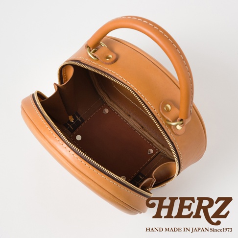 HERZ 品牌手工製作的棕色皮革圓形包，內部細節和金色拉鍊，下方有品牌 LOGO 和「HAND MADE IN JAPAN Since1973」字樣