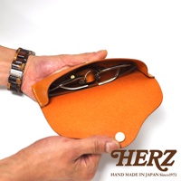 一只手拿著一副眼鏡，放入一個橘色皮革製的眼鏡收納盒。收納盒下方印有HERZ字樣和HAND MADE IN JAPAN Since1973。