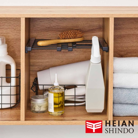 黑桿伸縮縫隙置物架，架上擺放著木柄梳子，下方收納著吹風機、髮油瓶、噴霧瓶，以及置物籃內的洗衣精瓶，展示於木紋層架空間。