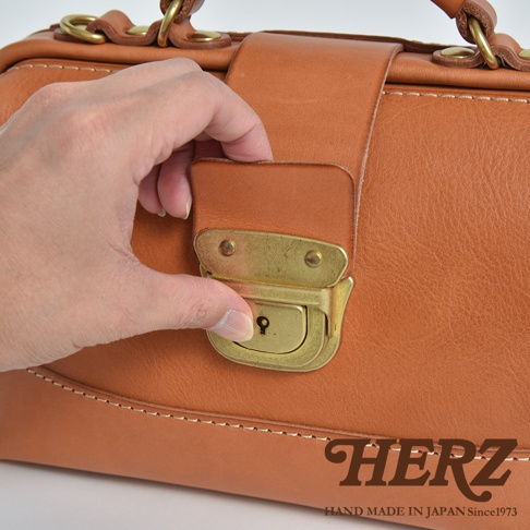 一隻手正輕輕按壓日本HERZ手工皮革迷你口金包的黃銅鎖扣，展現皮革溫潤的質感與細膩的縫線細節，圖片右下角印有品牌名稱與手工製造資訊。