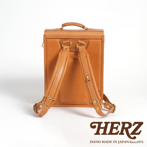 職人手作皮革小學生書包，採用天然皮革製成，呈現溫潤的焦糖棕色。書包設計簡潔，配有兩個可調節的皮革背帶，以及頂部提把，整體風格復古且實用。