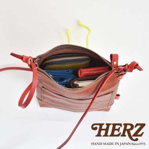 職人手作皮革直式隨身單肩包，呈現棕褐色牛皮材質，包內裝有綠色皮夾、紅色皮夾、藍色布料及筆記本，附有紅色皮革背帶，拉鍊為金色。