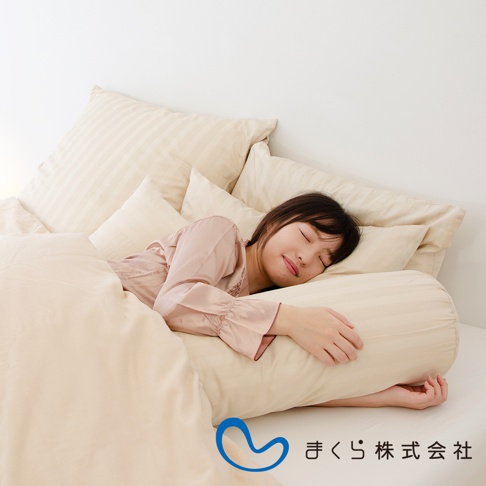 一位女子在床上枕著圓筒狀的米色條紋枕頭，周圍有數個方形枕頭和米色床單，整體色調柔和。