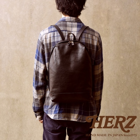 一位男性穿著藍灰色格紋襯衫，背著一個日本HERZ手工製作的深咖啡色皮革後背包，背包採用簡約的圓弧造型與柔軟皮革材質，展現職人手工藝質感。