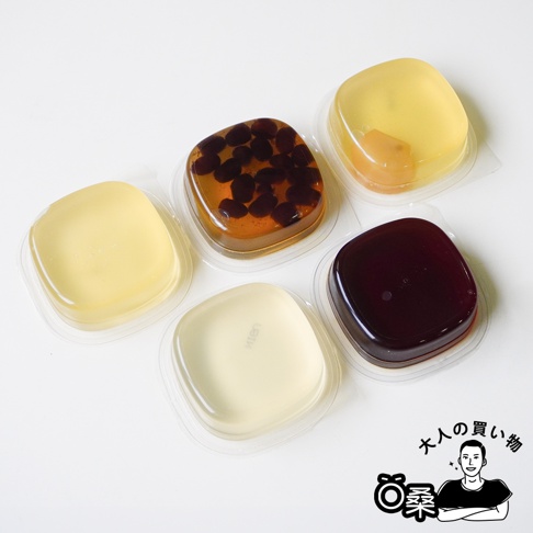 五個透明塑膠盒盛裝的日式甜點，包含紅豆口味、栗子口味、以及三款呈現淡雅半透明質感的葛餅，整體採俯視角度拍攝，右下角配有一位卡通人物的簡約線條插畫與大人的購物文字。
