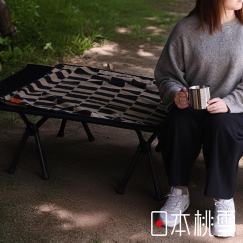 日本桃雪戶外格紋棉柔浴巾，上面鋪在黑色摺疊床架上，有人手持金屬杯坐在床邊。