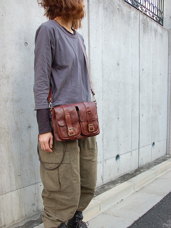 一位模特兒身穿灰色上衣和卡其色工裝褲，斜挎著一個棕色皮革雙口袋肩背包，背包上有兩個翻蓋和金色搭扣。
