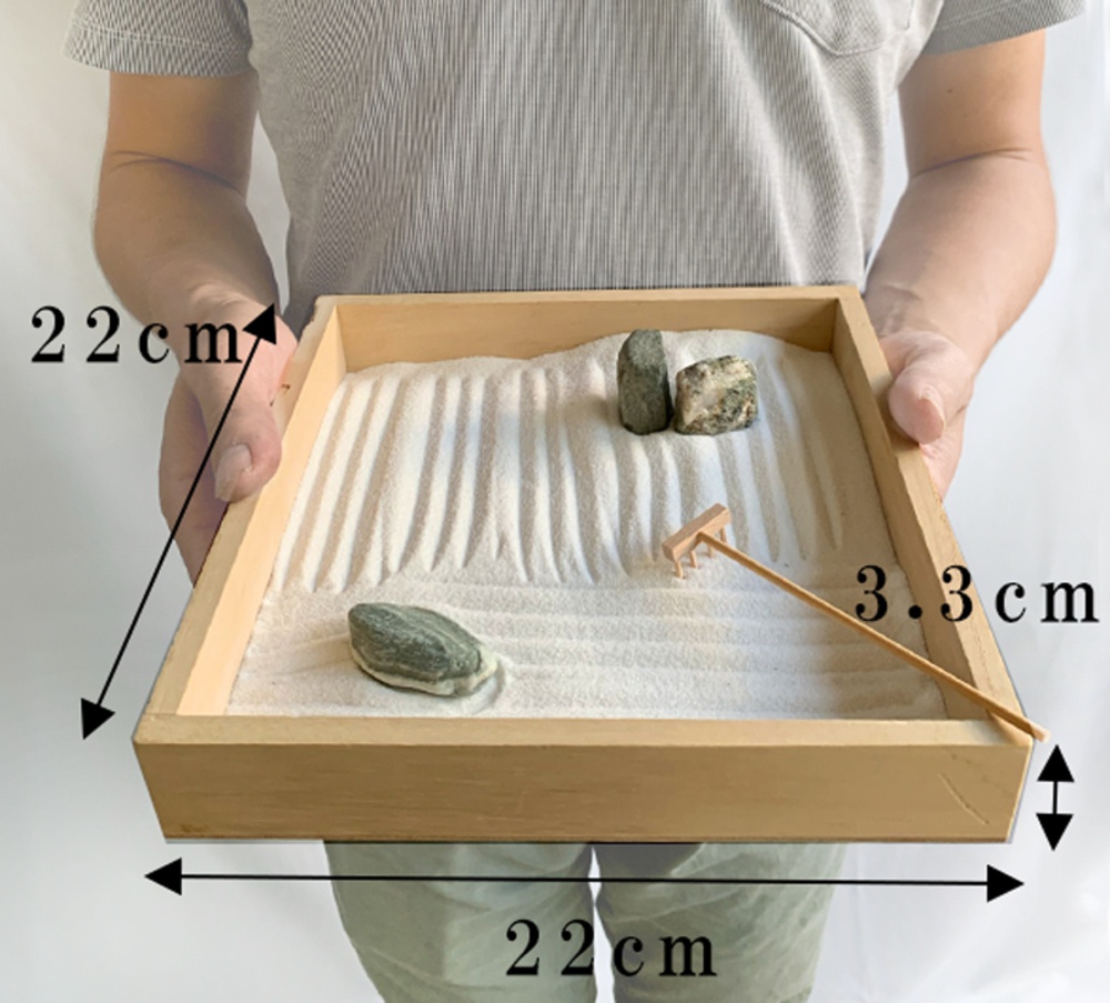 日本枯山水模型，木質淺盤，填充白色細沙，表面梳有波浪紋路，盤中置有兩塊灰綠色石頭及一把微型木耙。模型尺寸約為22x22x3.3公分。
